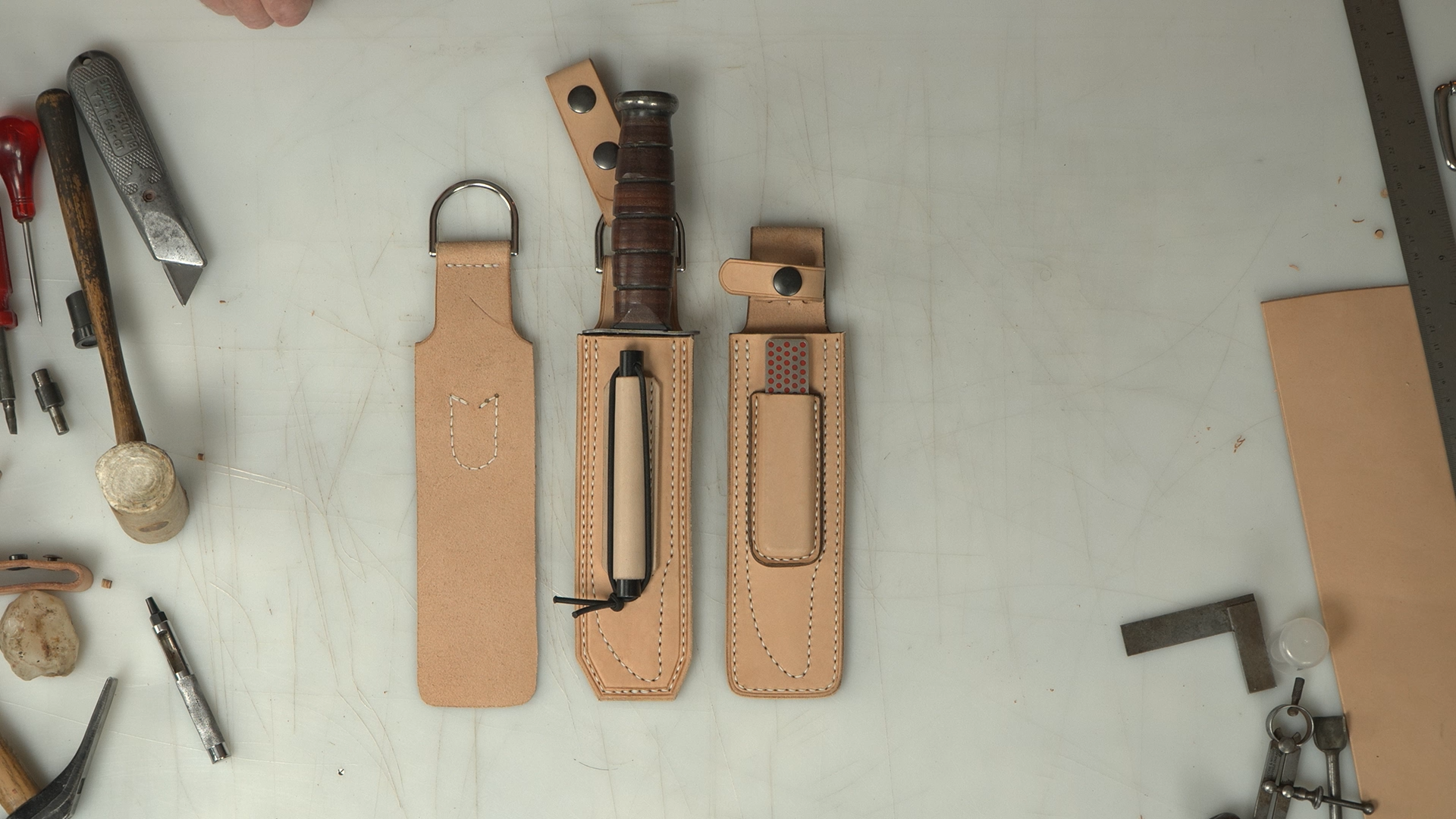 Three leather sheaths for knives on a light gray surface with tools in the background.