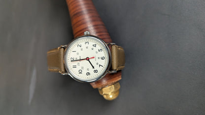 A vintage-style wristwatch with a brown leather strap and a white dial, resting on a wooden prop against a dark gray background.