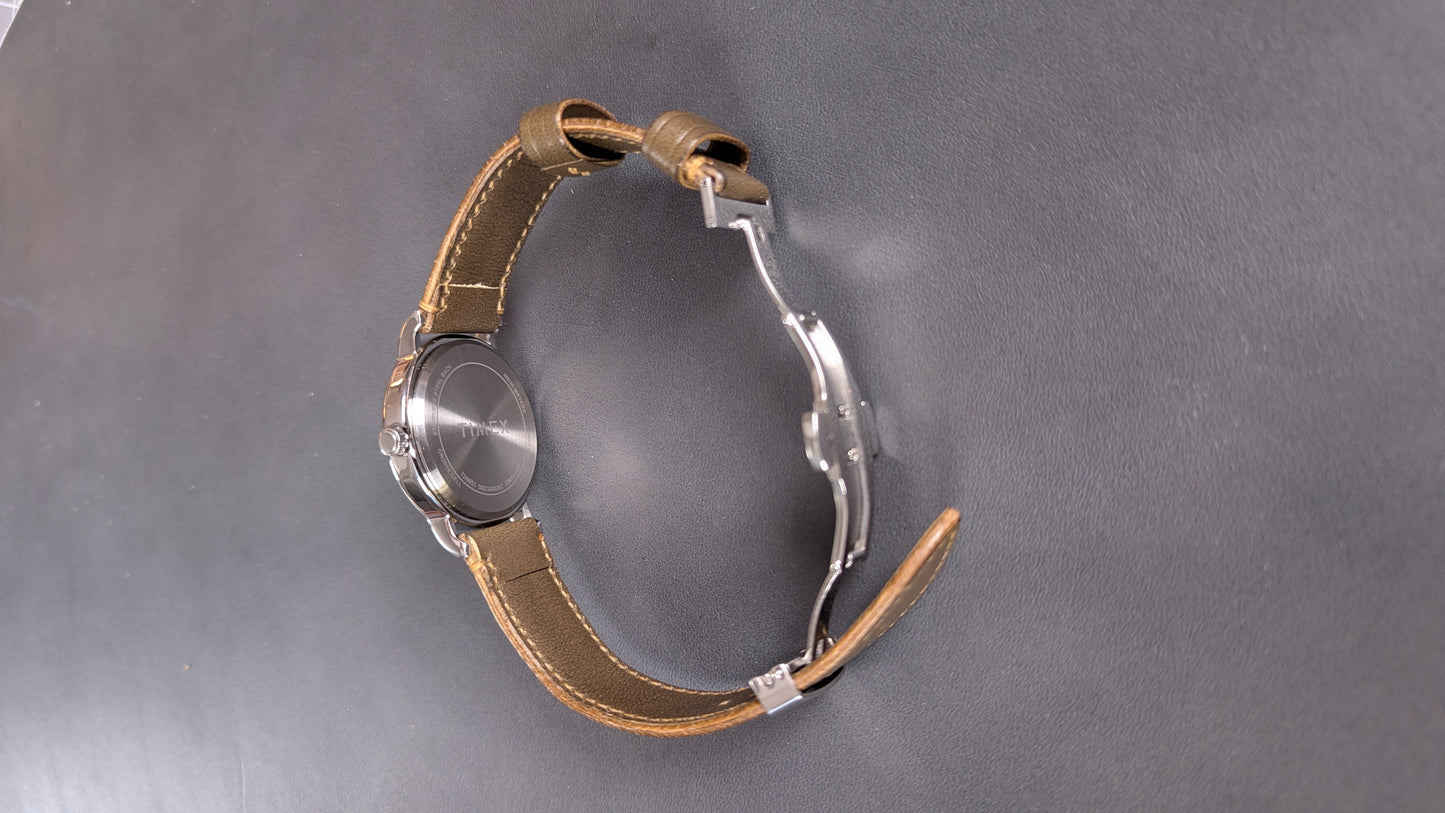 A vintage-style wristwatch with a round silver-tone case, a tan leather strap, and a distinctive silver metal buckle, photographed on a dark gray textured surface.