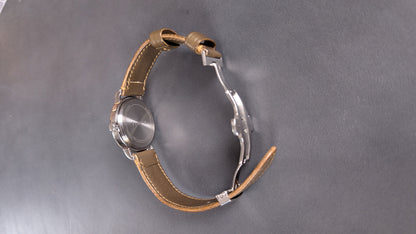 A vintage-style wristwatch with a round silver-tone case, a tan leather strap, and a distinctive silver metal buckle, photographed on a dark gray textured surface.